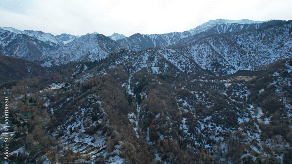 High snowy peaks stand in the distance. Drone view of the highway with cars, forest trails, blue sky with clouds. In places there is snow, a river runs. Mountainous area near Medeo, Almaty, Kazakhstan High snowy peaks stand in the distance. Drone view of the highway with cars, forest trails, blue sky with clouds. In places there is snow, a river runs. Mountainous area near Medeo, Almaty, Kazakhstan