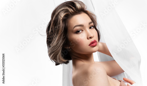 Alluring asian girl wrapped in waving white curtain with perfect facial skin, fresh spring make up looking at camera. Rejuvenation, skincare treatment concept.
