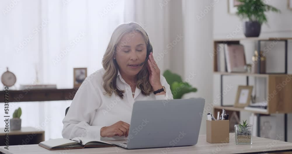 Freelance mature female call center agent wearing a headset and answering calls working on a laptop. One content customer service worker talking on a call and writing in notebook working remotely Freelance mature female call center agent wearing a headset and answering calls working on a laptop. One content customer service worker talking on a call and writing in notebook working remotely
