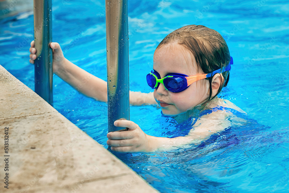 a little girl swims in an outdoor pool with blue clear water in the hot season, she is wearing a bright bathing suit and waterproof goggles