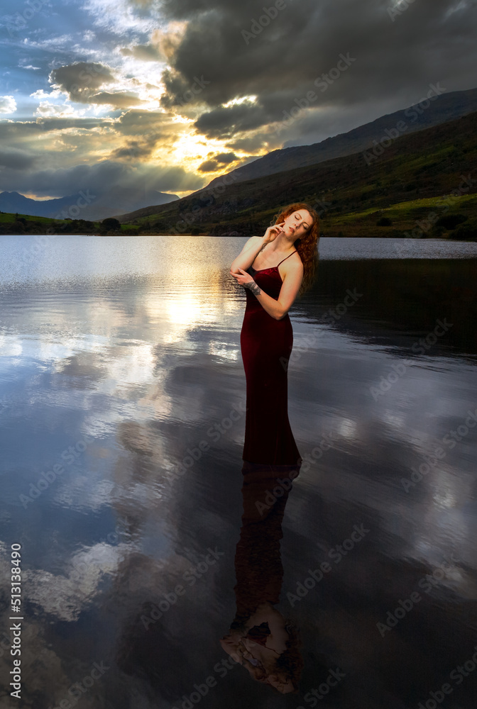 Fototapeta premium Woman with long red hair standing in the still water of a lake with a dramatic cloudy sunset