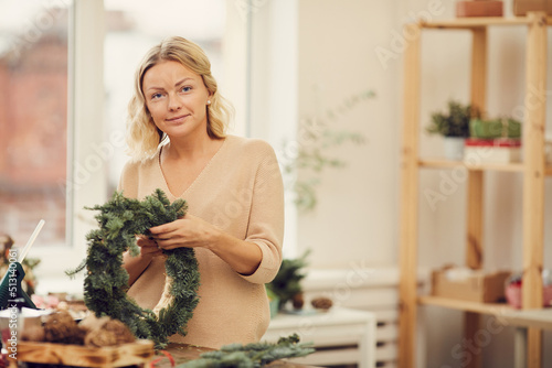 Portrait of smiling attractive young blonde woman in sweater standing at table in workshop and adding tree twigs to wreath
