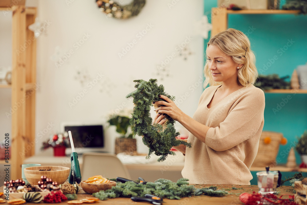 Smiling beautiful young woman with wavy hair standing at desk with messy decorations and checking base of Christmas wreath