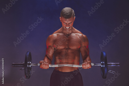 Brutal sweaty strong young man athlete with naked upper body standing doing workout with barbell and showing strong pumped up biceps over smoky background. Sport men body concept © zamuruev