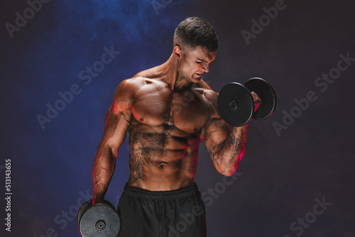 Brutal sweaty strong young man athlete with naked upper body standing doing workout with dumbbels and showing strong pumped up biceps over smoky background. Sport men body concept