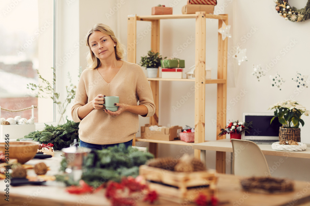 Portrait of content attractive blonde woman standing in cozy Christmas workshop on wreath production and drinking coffee Portrait of content attractive blonde woman standing in cozy Christmas workshop on wreath production and drinking coffee