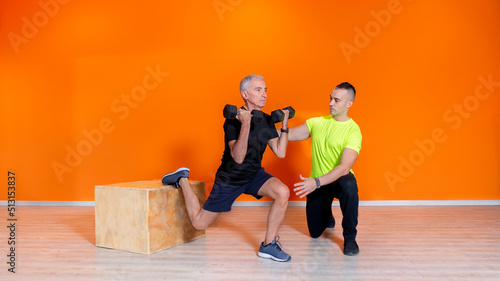 senior fit man doing weight lifting workout exercise standing at the gym isolated on orange background with pesonal trainer. mature people fitness training indoor. elderly person recovery concept photo