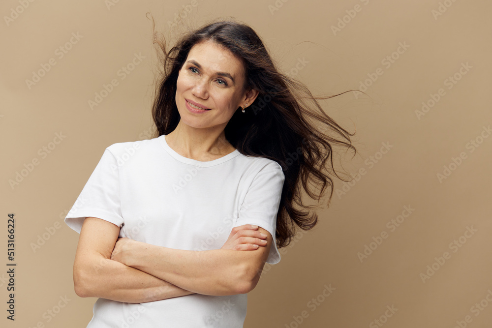 Fototapeta premium pleasant, funny woman with long well-groomed hair flying in the wind stands in a white T-shirt with her arms folded on her chest and smiles broadly with beautiful bright teeth with her eyes closed