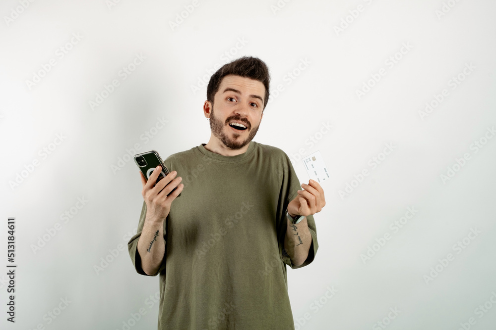 Caucasian young man wearing t-shirt posing isolated over white background holding credit or debit card and smartphone in his hands, free space. Caucasian young man wearing t-shirt posing isolated over white background holding credit or debit card and smartphone in his hands, free space.