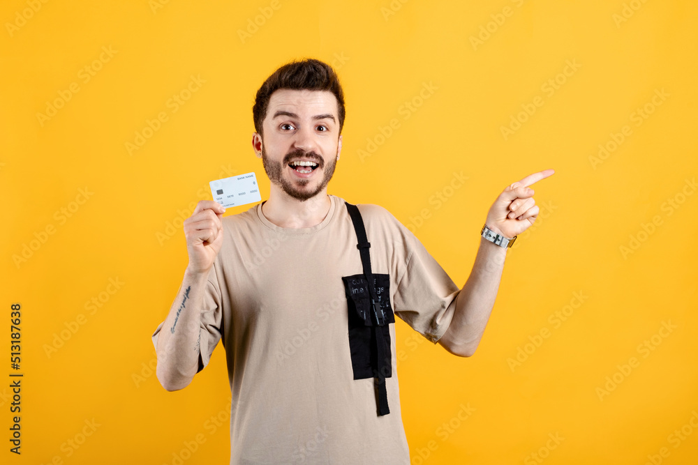 Cheerful young man wearing t-shirt posing isolated over yellow background holding credit bank card and pointing index finger aside. Shopping and finance concept. Cheerful young man wearing t-shirt posing isolated over yellow background holding credit bank card and pointing index finger aside. Shopping and finance concept.