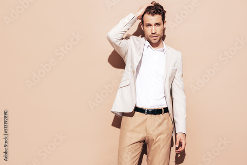 Portrait of handsome confident stylish hipster lambersexual model. Sexy modern man dressed in white elegant suit. Fashion male posing in studio near beige wall
