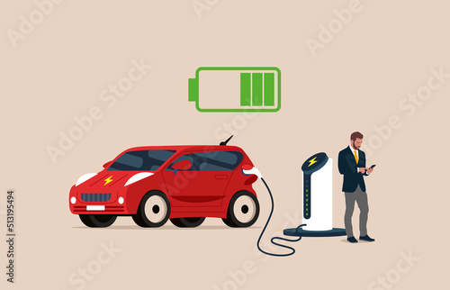 Handsome businessman plays smartphone while waiting for his EV Car or Electric charging at the charger station with the power cable supply plugged in.