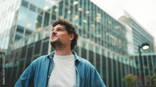 Close-up, man in denim shirt walks down the street, stops and looks around at the upper floors of modern buildings