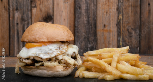 Fast food. Closeup view of a burger with crispy onions, provolone cheese, a grilled egg and fried potatoes, on the table with a wooden background. 	