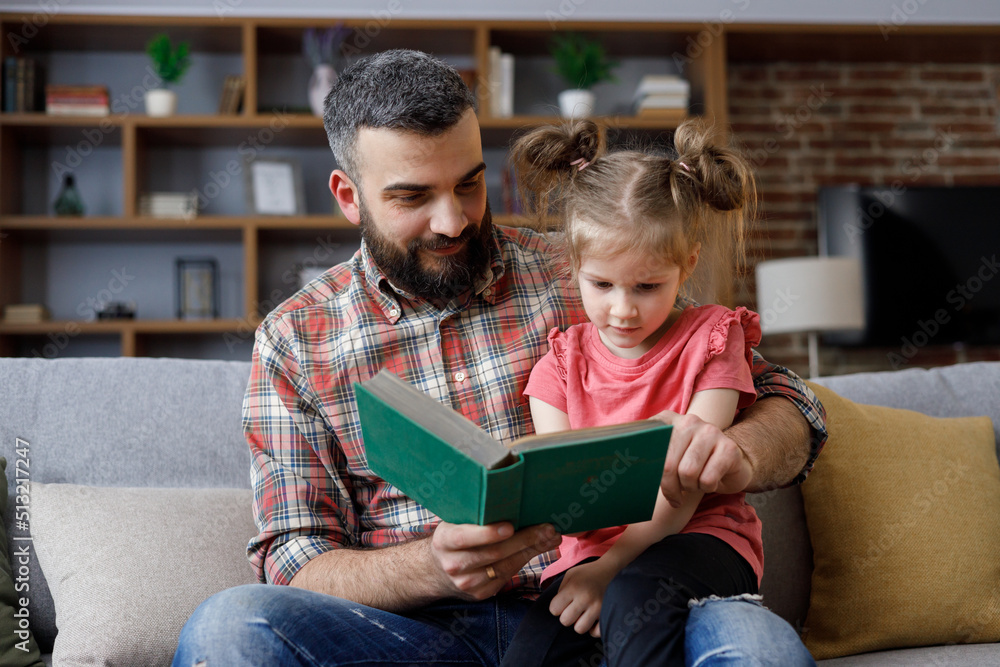 Young father and daughter read intersting book at home. Loving parent bearded man teaches his preschool child to read. Daddy reading fairy tale story. Parenthood, love and happy family concept Young father and daughter read intersting book at home. Loving parent bearded man teaches his preschool child to read. Daddy reading fairy tale story. Parenthood, love and happy family concept