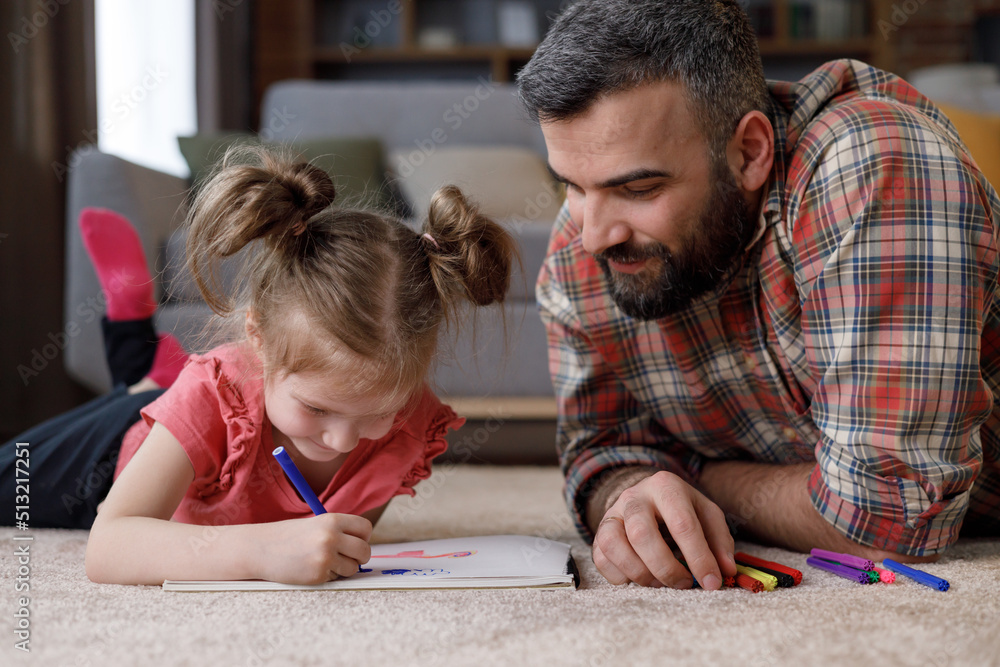 Young handsome father and cute daughter playing together. Dad and child are painting picture lying on the carpet near the sofa at home. Enjoy spending time together, home leisure. Father's day