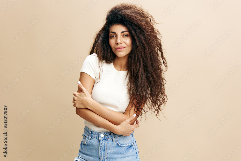 Gorgeous confident curly Latin female look at camera, crossing hands on the chest. Copy space for clothing fashion brands, free place for your ad. Studio shoot isolated over pastel beige background - obrazy, fototapety, plakaty 