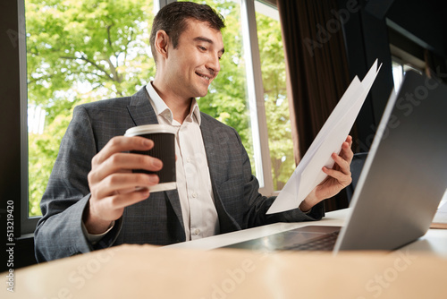Cheerful clerical worker checking documents while picking coffee cup