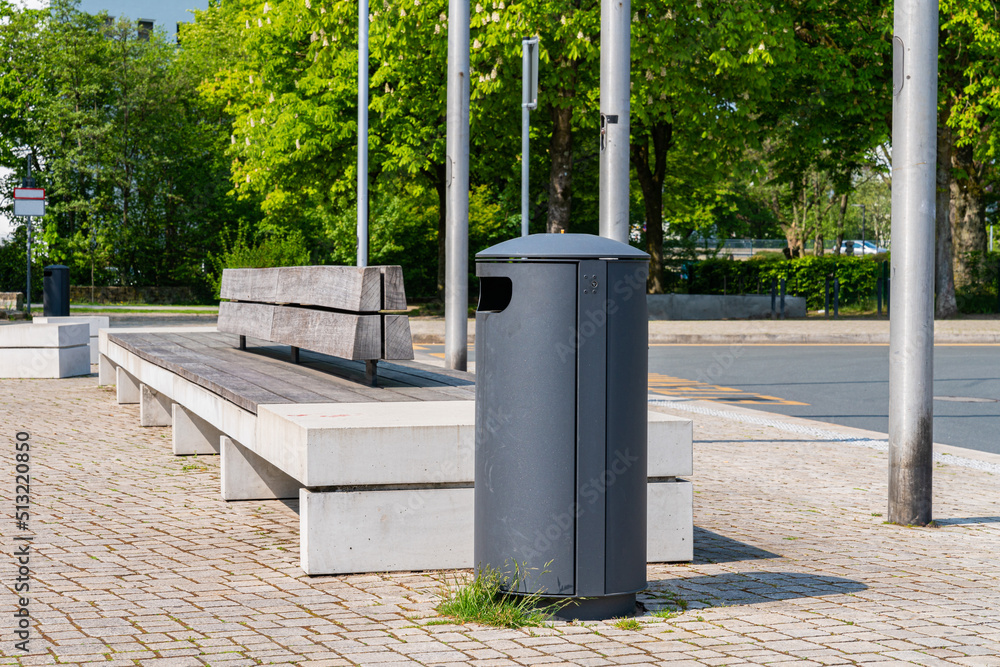 A metal trash bin and a long bench in a city park. A metal trash bin and a long bench in a city park.