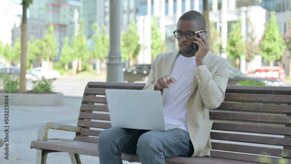 African Man Talking on Phone and using Laptop while Sitting Outdoor on Bench African Man Talking on Phone and using Laptop while Sitting Outdoor on Bench