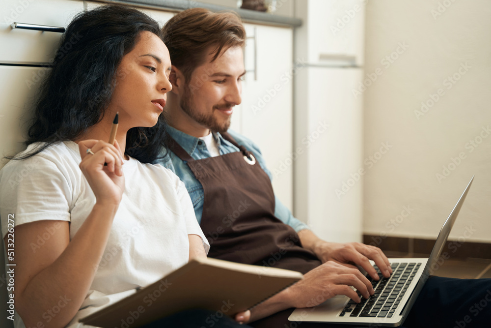 Pensive lady seated on kitchen floor beside male with computer Pensive lady seated on kitchen floor beside male with computer