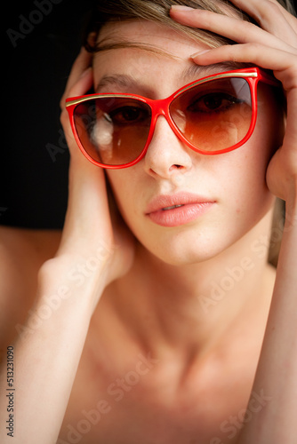 A female model in natural makeup wearing red sunglasses. Studio close head and shoulder portrait of a beautiful young model set against black.