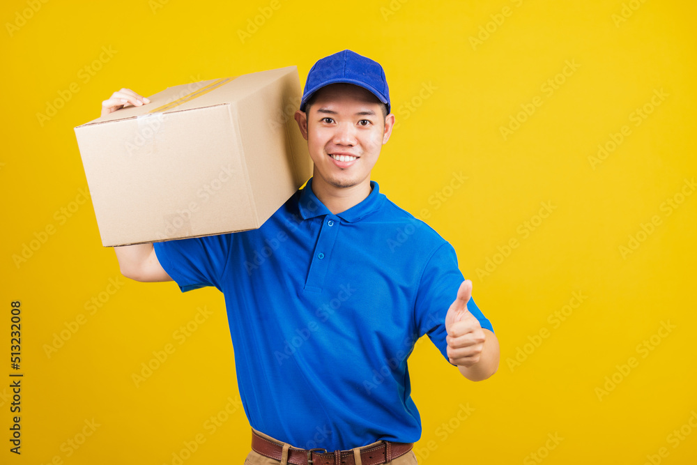 Portrait excited delivery happy man logistic standing he smile wearing blue t-shirt and cap uniform holding parcel box show thumb up finger looking to camera, studio shot isolated on yellow background Portrait excited delivery happy man logistic standing he smile wearing blue t-shirt and cap uniform holding parcel box show thumb up finger looking to camera, studio shot isolated on yellow background