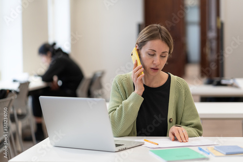 Middle-aged female busy with documents talking on cellphone, working on laptop in campus open space. Focused mature student woman studying preparing for exam in university. Education has no age limit