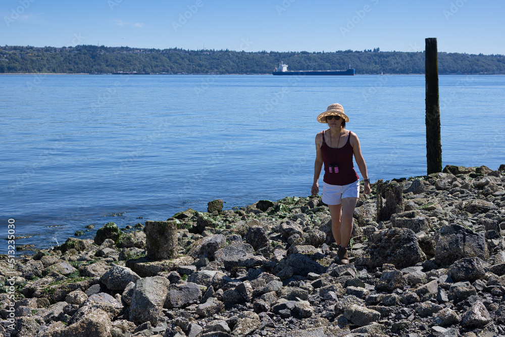 A woman wearing white shorts, a tank top, sunglasses, and a summer hat walking on a rocky beach in Tacoma, Washington.