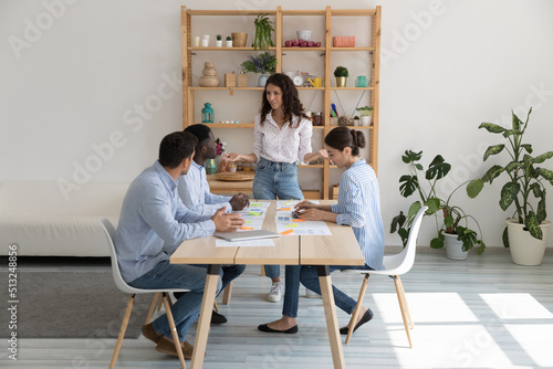 Hispanic team leader makes speech standing in front of seated at desk staff or company clients  businesspeople take part in group meeting in office. Teamwork  sales statistics analysis process concept