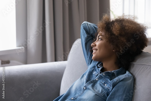 Young African 18s girl rest leaned on couch staring into distance looks serene enjoy carefree weekend leisure alone at home, daydream, breath fresh conditioned air feels peaceful. Relaxation concept