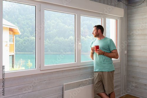 Relaxed man holding a mug and enjoying a lake views from the window of his apartment.