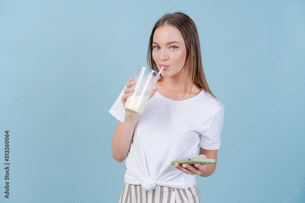 Girl stands with cocktails and smartphone on copy blue background in studio in light casual clothes and smiles sweetly. Photo gorgeous woman standing in white t-shirt shaking glass with milkshake Girl stands with cocktails and smartphone on copy blue background in studio in light casual clothes and smiles sweetly. Photo gorgeous woman standing in white t-shirt shaking glass with milkshake