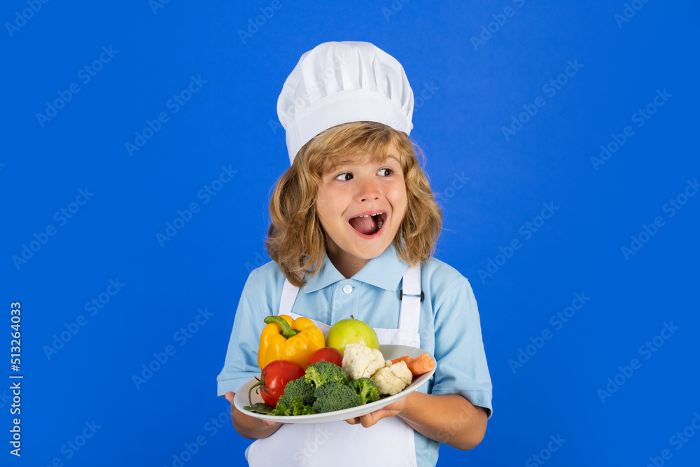 Chef kid boy making fresh vegetables for healthy eat. Portrait of little child in form of cook isolated on grey background. Kid chef hold plate with vegetables. Chef kid boy making fresh vegetables for healthy eat. Portrait of little child in form of cook isolated on grey background. Kid chef hold plate with vegetables.