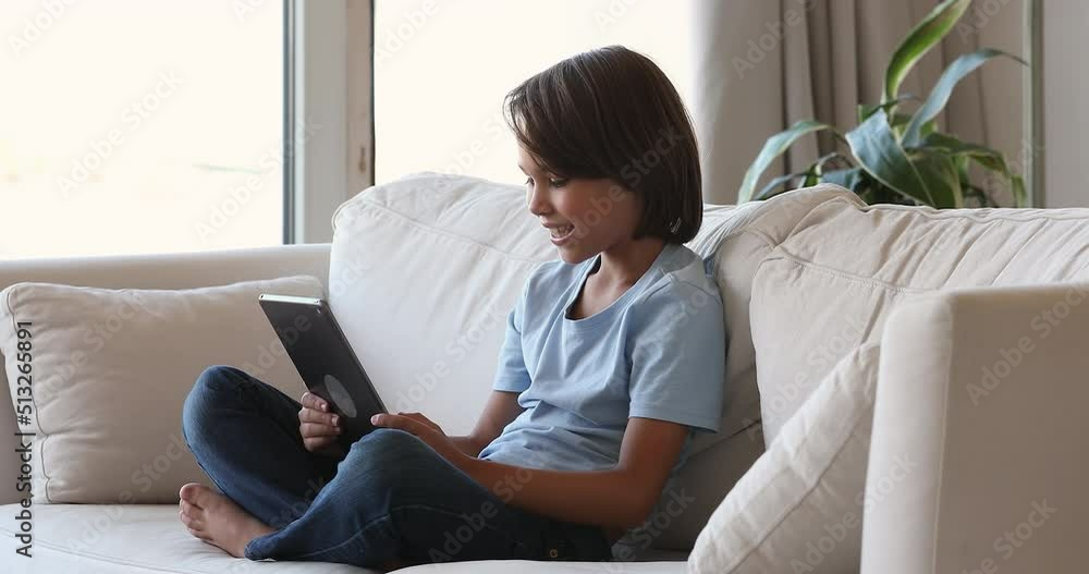 Happy engaged gen Z pre teen school boy playing virtual online video game on digital gadget, using tablet computer at home couch, touching screen, looking at display, smiling. Communication concept Happy engaged gen Z pre teen school boy playing virtual online video game on digital gadget, using tablet computer at home couch, touching screen, looking at display, smiling. Communication concept