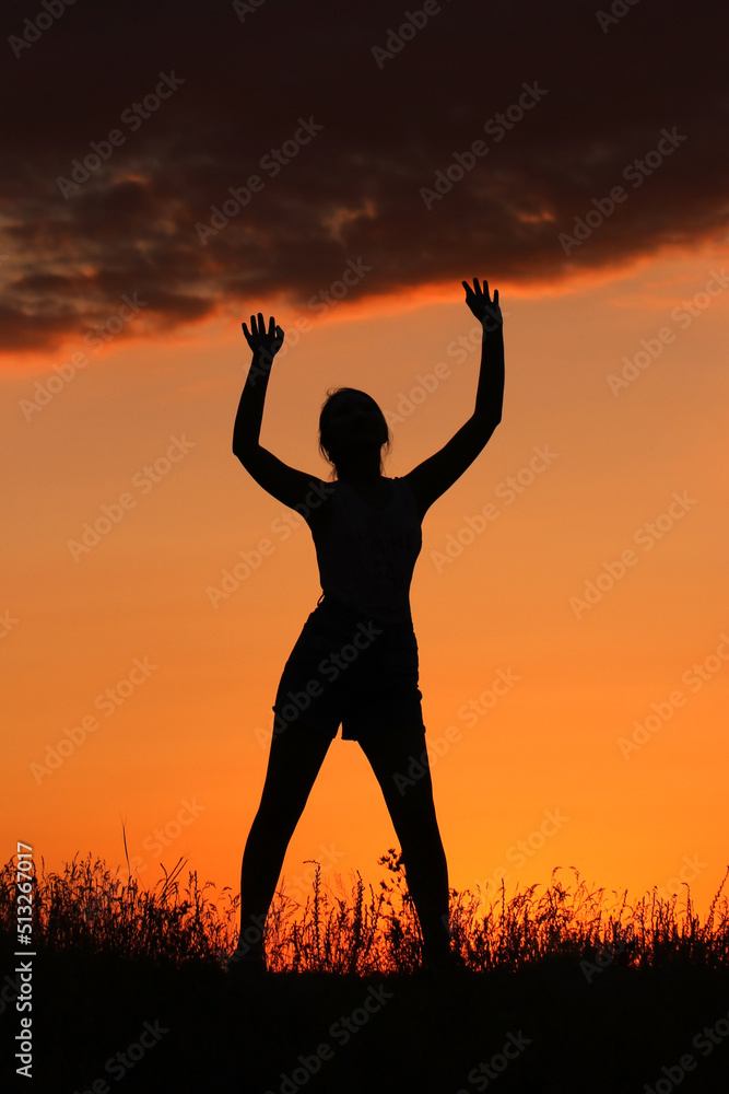 The silhouette of a confident girl stands against the background of the sunset and holds the clouds