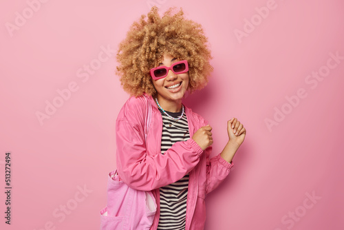 Carefree cheerful young woman wears sunglasses striped jumper and jacket carries fabric bag dances with rhythm of music isolated over pink background has good mood. People and happiness concept
