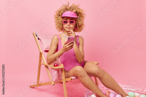 Thoughtful curly haired young woman wears sunglasses swimsuit and earrings holds mobile phone being deep in thoughts poses on deck chair enjoys summer resort has perfect holiday. Rest concept