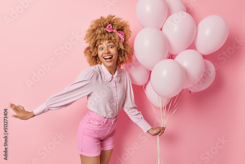 Positive surpised woman spends free time on welcome party holds bunch of inflated balloons wears blouse and shorts feels very happy dances against pink background. Birthday celebration concept