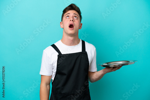 Young waitress with tray isolated on blue background looking up and with surprised expression