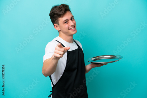Young waitress with tray isolated on blue background pointing front with happy expression photo