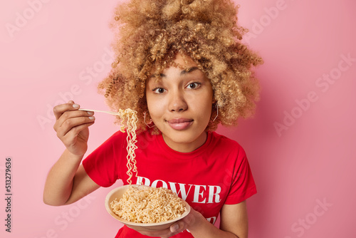 Fototapete Photo of serious curly haired young woman eats appetizing noodles has delicious lunch enjoys favorite meal dressed in casual red t shirt isolated over pink background