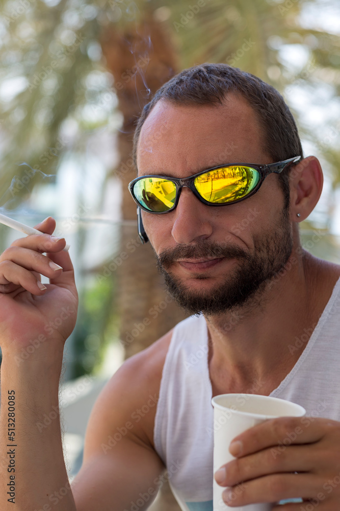Brutal caucasian beautiful guy smoking wearing sunglasses and white t-shirt  in a cafe. American man with a cigarette in his hand. Handsome Jewish man  mustache and beard drinking hot espresso coffee Stock