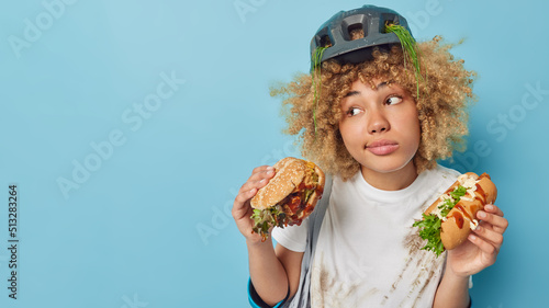 Horizontal shot of thoughtful female bicycle rider wears white dirty t shirt and protective helmet holds burger and hotdog eats unhealthy food isolated over blue background empty space for promo