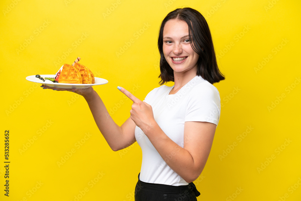 Young caucasian woman holding waffles isolated on yellow background and pointing it Young caucasian woman holding waffles isolated on yellow background and pointing it