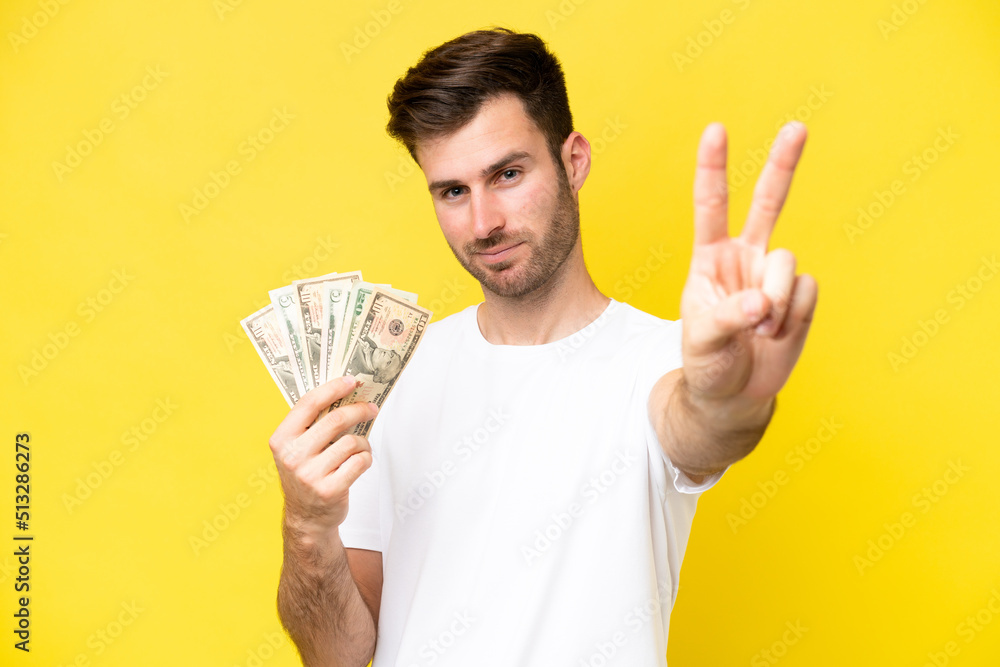 Young caucasian man taking a lot of money isolated on yellow background smiling and showing victory sign Young caucasian man taking a lot of money isolated on yellow background smiling and showing victory sign