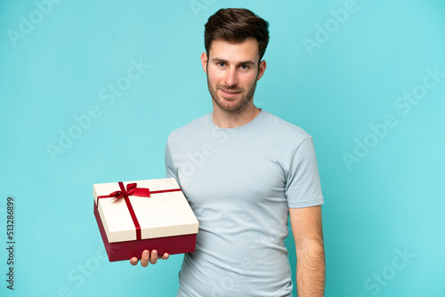 Young caucasian man holding a gift isolated on blue background smiling a lot