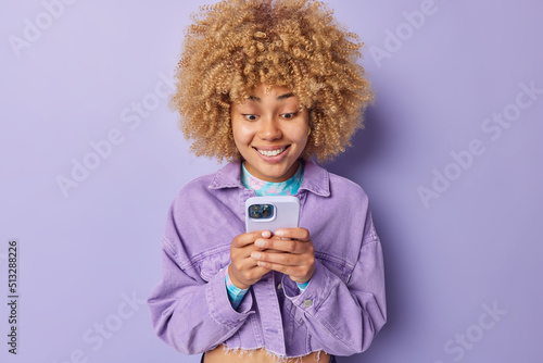 Fashionable happy young woman with curly hair dressed in stylish jacket uses mobile phone chats with friends checks emial or makes shopping or orders food online isolated over purple background