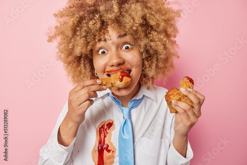 Harmful food concept. Curly haired woman eats delicious fried nuggets with appetite stares impressed at camera dressed in white formal shirt and blue tie poses indoor. Guilty pleasure concept