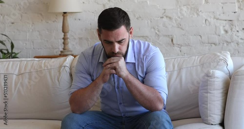 Stressed concerned millennial man thinking over bad news, problem solving, sitting on couch at home, touching, rubbing face, leaning chin on hands, feeling depressed, frustrated, coping with crisis